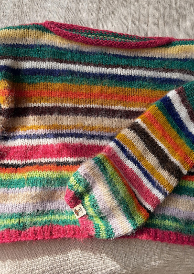 May include: A colorful, striped sweater with a matching sleeve. The sweater features horizontal bands of various colors, including pink, green, orange, and brown. The neckline and sleeve cuff are trimmed with a bright pink. The sweater is made of a soft, textured yarn.