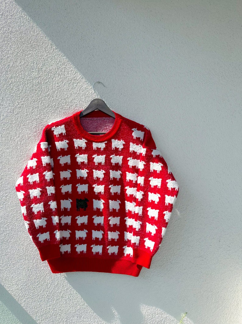 Może przedstawiać: Czerwony sweter z wzorem białych owiec. Jedna czarna owca wyr&oacute;żnia się wśr&oacute;d białych owiec.