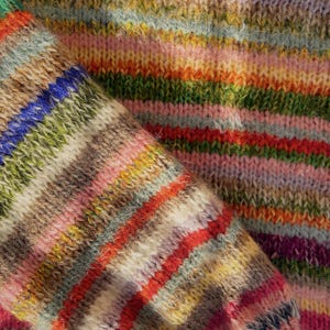 May include: Close-up of a knitted garment with a vibrant, multicolored striped pattern. The design features horizontal bands of various colors, including red, orange, green, blue, and pink. The texture of the knitwear is visible, suggesting a soft and cozy material.
