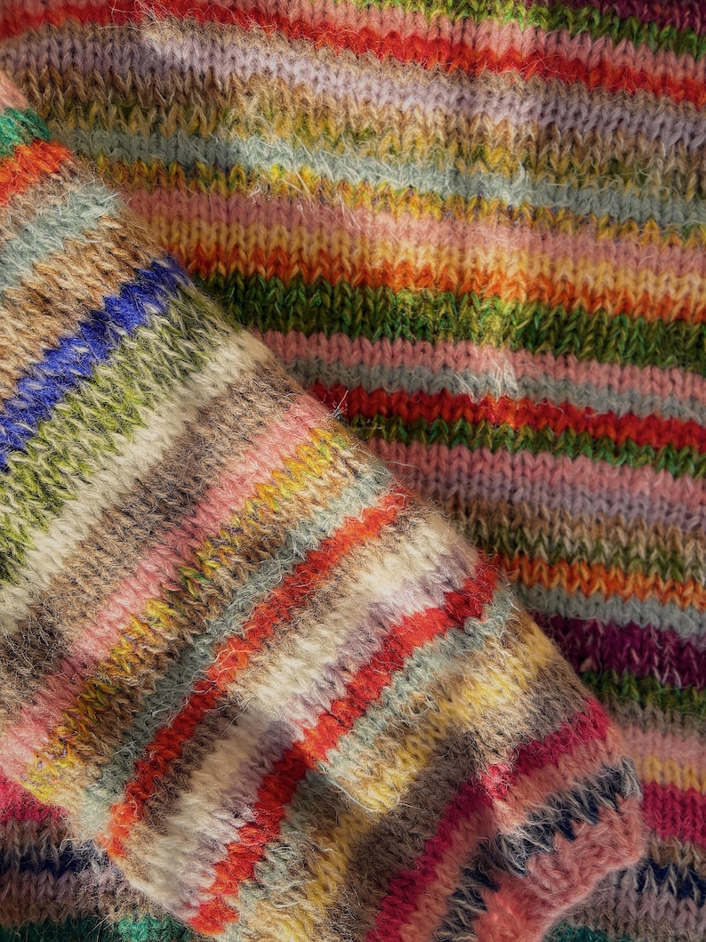 May include: Close-up of a knitted garment with a vibrant, multicolored striped pattern. The design features horizontal bands of various colors, including red, orange, green, blue, and pink. The texture of the knitwear is visible, suggesting a soft and cozy material.