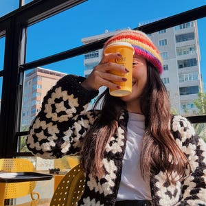 May include: A person holding a yellow coffee cup with the word "coffee" printed on it. They are wearing a rainbow striped beanie, a black and white crochet cardigan, and a white shirt. The background shows a blue sky and buildings.