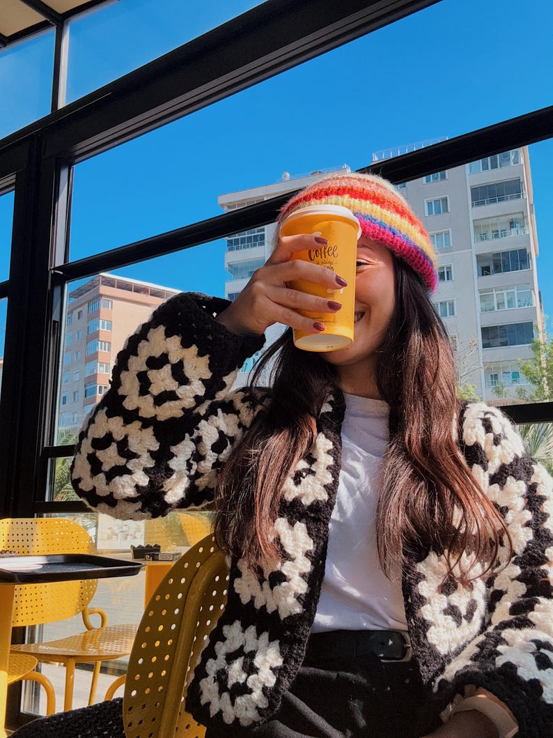 May include: A person holding a yellow coffee cup with the word "coffee" printed on it. They are wearing a rainbow striped beanie, a black and white crochet cardigan, and a white shirt. The background shows a blue sky and buildings.
