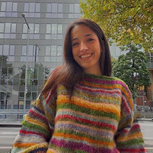 May include: A colorful, striped sweater in shades of green, orange, pink, and purple. The sweater has a round neckline and long sleeves. The background includes a building with many windows and a tree.