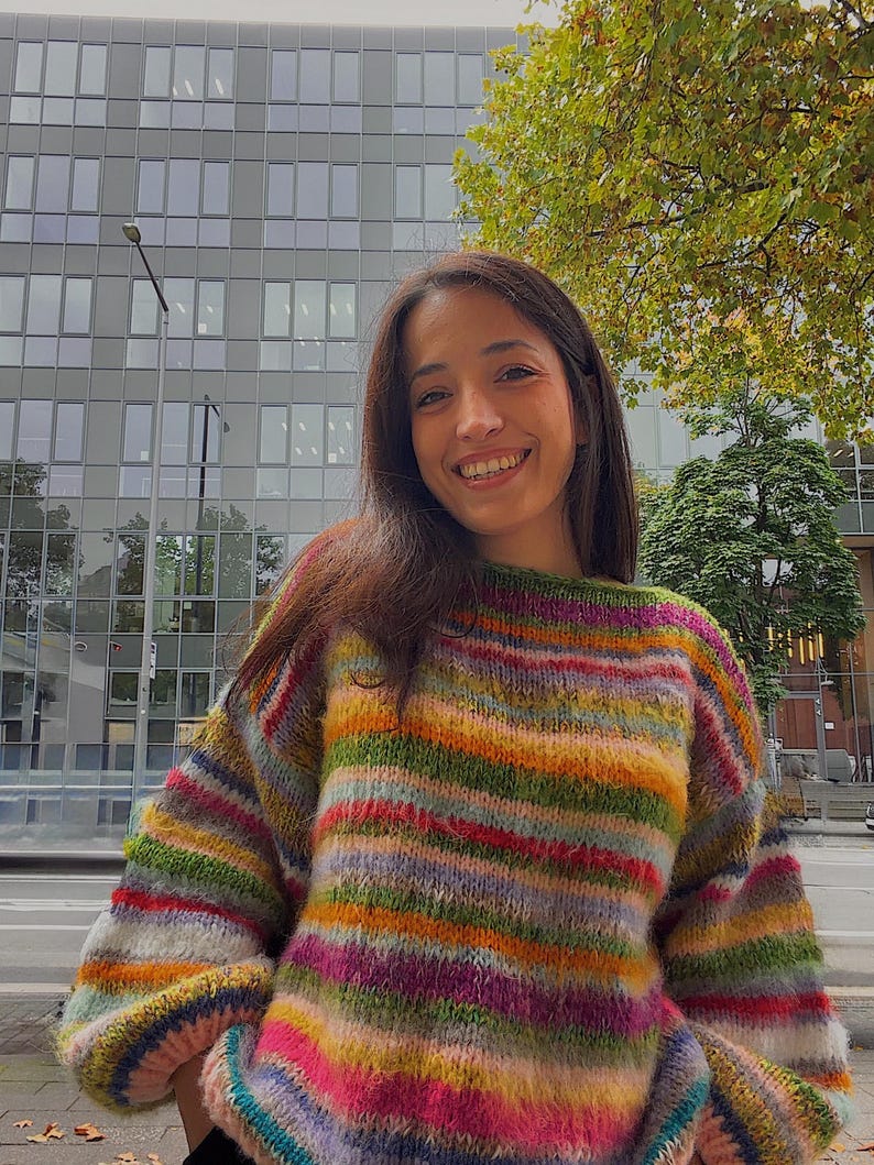 May include: A colorful, striped sweater in shades of green, orange, pink, and purple. The sweater has a round neckline and long sleeves. The background includes a building with many windows and a tree.