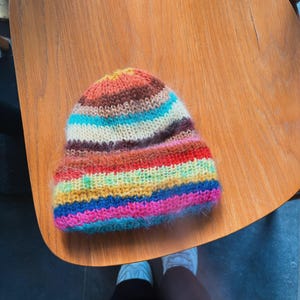 May include: A colorful, hand-knitted beanie hat with horizontal stripes in shades of orange, brown, blue, white, red, yellow, and pink. The hat is made of a soft, fuzzy yarn and is sitting on a wooden surface.