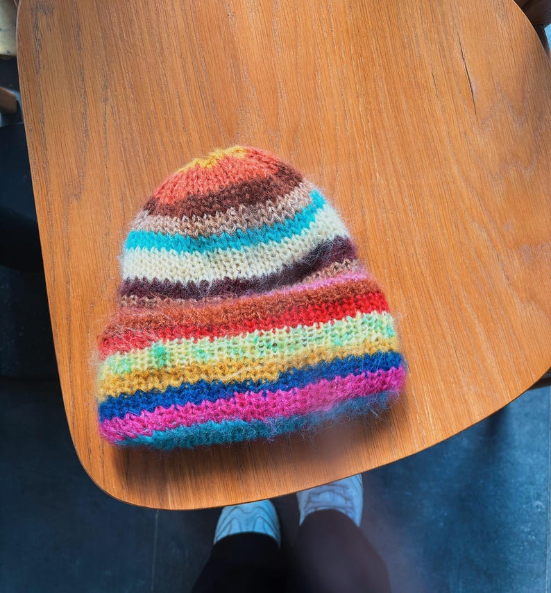 May include: A colorful, hand-knitted beanie hat with horizontal stripes in shades of orange, brown, blue, white, red, yellow, and pink. The hat is made of a soft, fuzzy yarn and is sitting on a wooden surface.