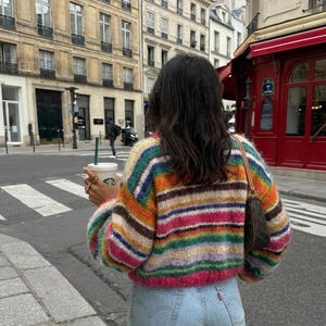 May include: A cropped, multi-colored striped sweater with a round neckline. The sweater features horizontal stripes in shades of pink, orange, yellow, green, and blue. The person is wearing light blue jeans and carrying a coffee cup.