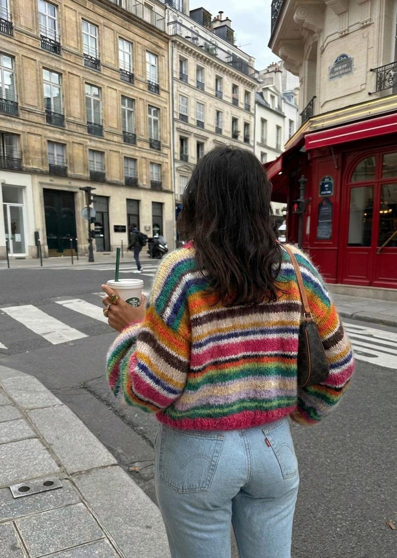 May include: A cropped, multi-colored striped sweater with a round neckline. The sweater features horizontal stripes in shades of pink, orange, yellow, green, and blue. The person is wearing light blue jeans and carrying a coffee cup.