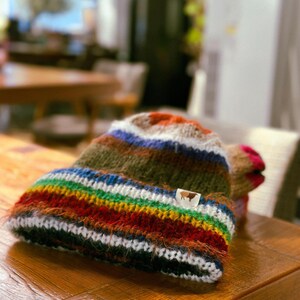 May include: A colorful, hand-knitted beanie hat with a rainbow striped brim and a brown and white tag. The hat features a mix of colors including green, blue, red, and orange. The hat is made of a soft, textured yarn and is displayed on a wooden surface.