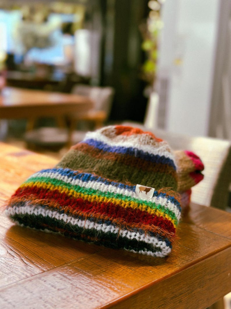 May include: A colorful, hand-knitted beanie hat with a rainbow striped brim and a brown and white tag. The hat features a mix of colors including green, blue, red, and orange. The hat is made of a soft, textured yarn and is displayed on a wooden surface.