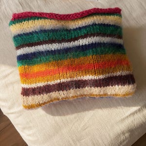 May include: A colorful, hand-knitted cowl featuring horizontal stripes in various colors, including green, yellow, orange, and brown. The top edge is a vibrant pink. The cowl is made of soft yarn and appears cozy and warm.