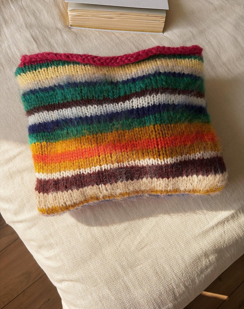 May include: A colorful, hand-knitted cowl featuring horizontal stripes in various colors, including green, yellow, orange, and brown. The top edge is a vibrant pink. The cowl is made of soft yarn and appears cozy and warm.