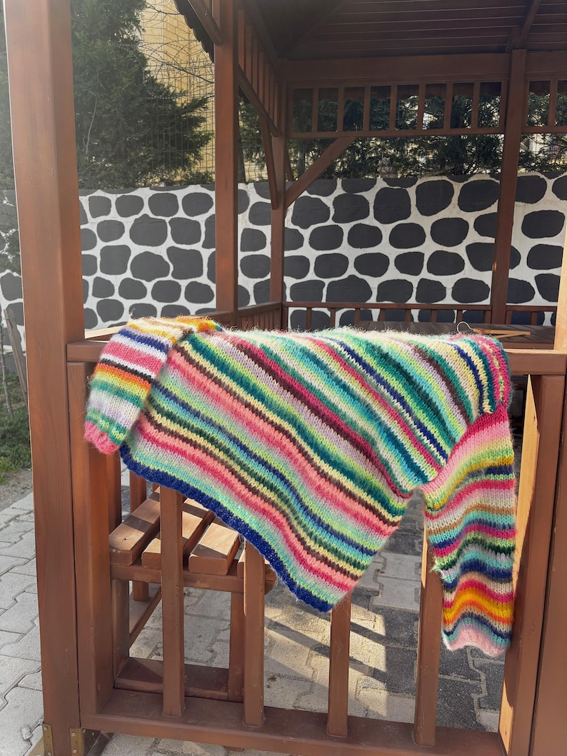 Może przedstawiać: Kolorowy, prążkowany sweter z długimi rękawami. Sweter jest zawieszony na drewnianej balustradzie.