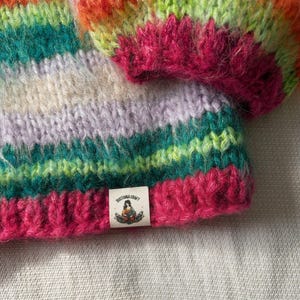 May include: A close-up of a hand-knitted hat with a vibrant, multicolored striped pattern. The hat features bands of pink, green, orange, and purple. A small tag with the text "SUSTANA CRAFT" and a graphic is sewn on the brim.