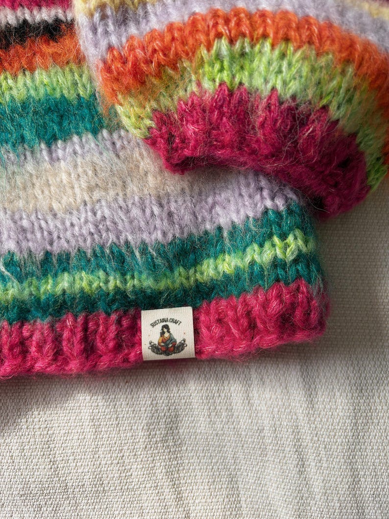 May include: A close-up of a hand-knitted hat with a vibrant, multicolored striped pattern. The hat features bands of pink, green, orange, and purple. A small tag with the text "SUSTANA CRAFT" and a graphic is sewn on the brim.