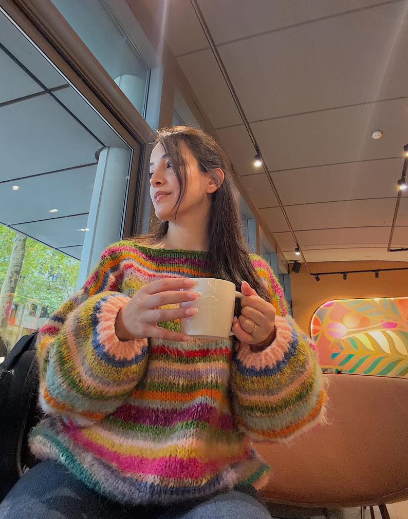 May include: A colorful, striped sweater with a boat neck and long sleeves. The sweater features a mix of pink, green, blue, and yellow stripes. A white mug is held in the hands.