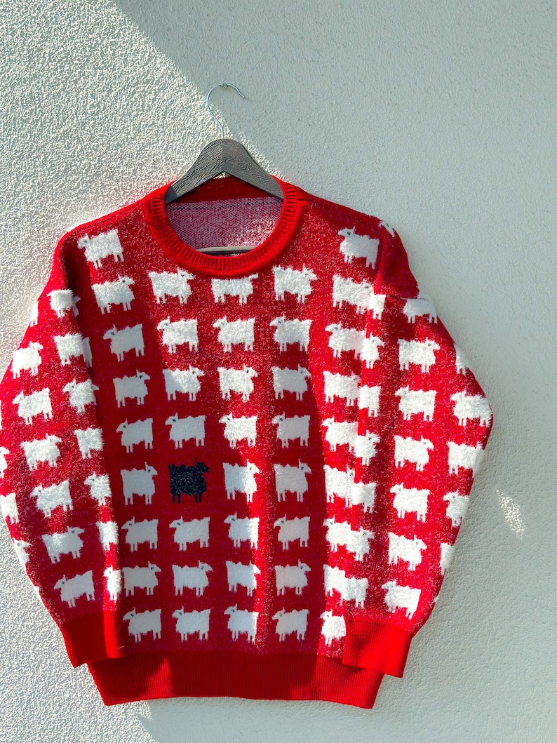Może przedstawiać: Czerwony sweter z wzorem białych owiec. Jedna czarna owca wyr&oacute;żnia się w środku swetra.