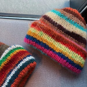 May include: Two colorful, striped knit beanies. The beanies feature horizontal stripes in shades of brown, orange, red, yellow, green, blue, and pink. The hats are made of a soft, fuzzy yarn.