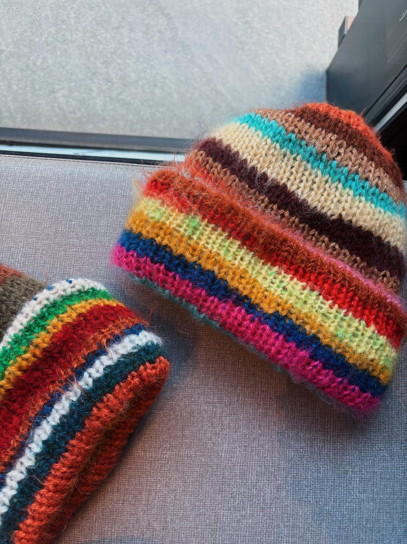 May include: Two colorful, striped knit beanies. The beanies feature horizontal stripes in shades of brown, orange, red, yellow, green, blue, and pink. The hats are made of a soft, fuzzy yarn.