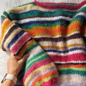 May include: A colorful, hand-knitted sweater with horizontal stripes in various colors, including pink, green, blue, and orange. The sweater has a round neckline with a pink trim and long sleeves. The yarn appears soft and fluffy.