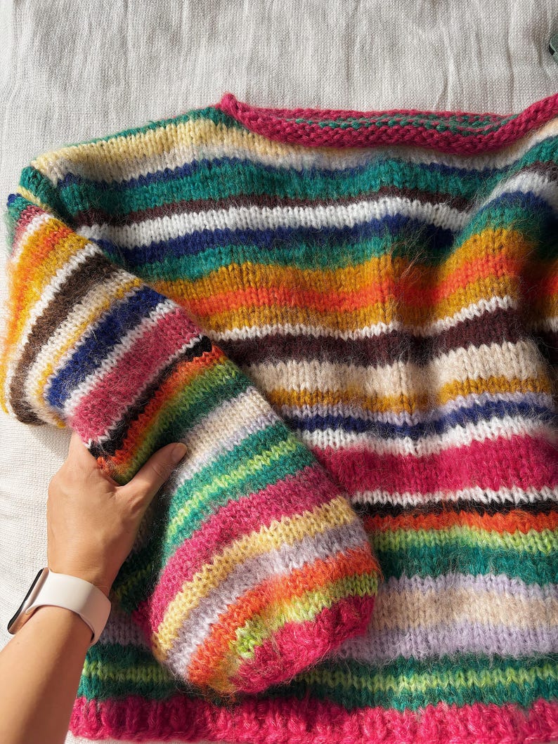 May include: A colorful, hand-knitted sweater with horizontal stripes in various colors, including pink, green, blue, and orange. The sweater has a round neckline with a pink trim and long sleeves. The yarn appears soft and fluffy.