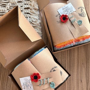 May include: Open cardboard boxes containing handcrafted items. Each box holds a folded textile, a red crocheted flower, and a set of crocheted rings. The boxes are lined with brown paper and include a thank you card.