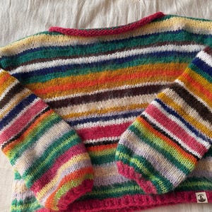 May include: A colorful, striped sweater with long sleeves. The sweater features a vibrant mix of colors including pink, green, yellow, and brown. The neckline and cuffs are trimmed in a bright pink. A book is visible in the background.