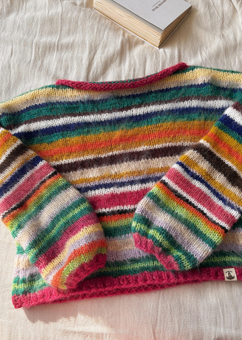 May include: A colorful, striped sweater with long sleeves. The sweater features a vibrant mix of colors including pink, green, yellow, and brown. The neckline and cuffs are trimmed in a bright pink. A book is visible in the background.