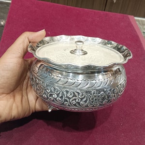 May include: A silver bowl with a lid, featuring intricate floral designs and three feet. The bowl has a scalloped edge and a smooth, polished finish.
