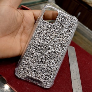 May include: A clear phone case with a silver, intricate floral design. The design is raised and textured, giving the case a 3D effect.