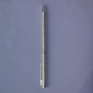 Puede incluir: Un bol&iacute;grafo de metal en tono plateado con un agarre texturizado de patr&oacute;n de diamantes a lo largo del cuerpo. El bol&iacute;grafo incluye una punta c&oacute;nica lisa y un clip de bolsillo recto y funcional unido a la parte superior del cuerpo cil&iacute;ndrico.