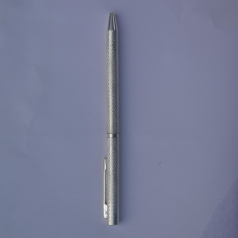 Puede incluir: Un bol&iacute;grafo de metal en tono plateado con un agarre texturizado de patr&oacute;n de diamantes a lo largo del cuerpo. El bol&iacute;grafo incluye una punta c&oacute;nica lisa y un clip de bolsillo recto y funcional unido a la parte superior del cuerpo cil&iacute;ndrico.