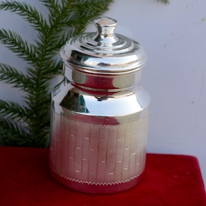 May include: A silver jar with a lid, featuring a decorative embossed pattern. The jar has a shiny, polished finish.