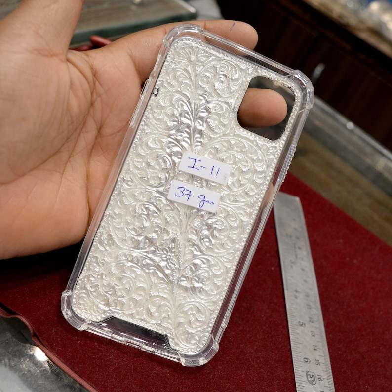 May include: A clear phone case with a silver, ornate design. The case has a label that reads "I-11 37 gm".