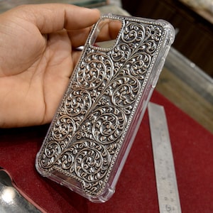 May include: A clear phone case with a silver, intricate floral design. The design is raised and textured, giving the case a 3D effect.