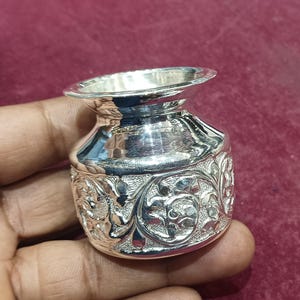 May include: A small, silver-colored pot with a wide, flared rim and a rounded body. The pot features intricate floral and leaf patterns around its base. The object is held in a hand against a burgundy background, highlighting its polished surface and detailed design.