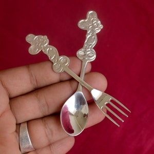 May include: A silver baby spoon and fork set with a cartoon character design. The spoon has a rounded bowl and the fork has three tines.