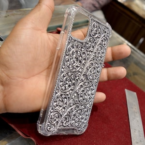 May include: A clear phone case with a silver, intricate floral design. The design is raised and has a textured finish.