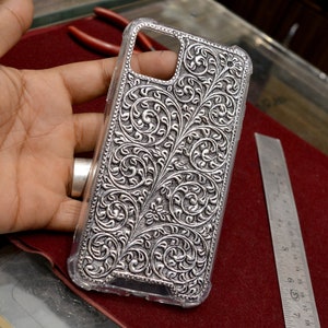 May include: A silver phone case with an intricate floral design. The case is made of metal and has a clear plastic back.