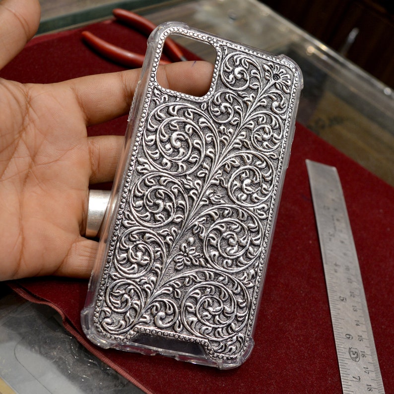 May include: A silver phone case with an intricate floral design. The case is made of metal and has a clear plastic back.