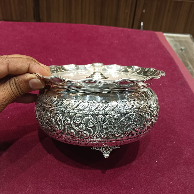 May include: A silver bowl with an ornate design and a lid. The bowl has three feet and is decorated with floral patterns.