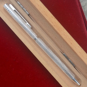 Puede incluir: Un bol&iacute;grafo de metal en tono plateado con un patr&oacute;n de diamantes texturizado y entrecruzado a lo largo del cuerpo y la tapa, equipado con un clip de bolsillo liso, presentado dentro de un estuche de madera clara con forro de terciopelo rojo.