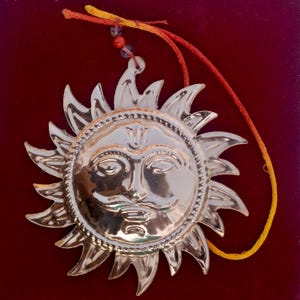 May include: A polished copper sun wall hanging featuring a central face with a moustache and stylised rays. The ornament includes a hanging loop with red and yellow threads and small clear and red beads, with the number 1986 engraved on the surface.
