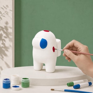May include: A white Among Us-style figurine with red and blue paint details. A hand is shown painting the figurine with a paintbrush. Paint pots and brushes are in the foreground. The figurine is being painted.