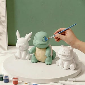 May include: Three unpainted ceramic figurines of cartoon creatures, including a white Pikachu, a white Bulbasaur, and a Squirtle being painted with a blue brush. The figures are arranged on a circular white base with small paint pots nearby.