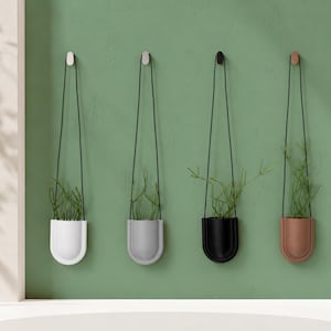 Op de afbeelding: Vier hangende plantenbakken in wit, grijs, zwart en koper, elk met een plant erin. De plantenbakken hangen aan de muur met een zwart koord en zijn op een groene muur gemonteerd.