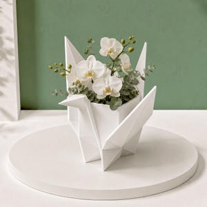 Może przedstawiać: Biała, geometryczna doniczka w kształcie żurawia origami, zawierająca bukiet białych storczyków i zielonych gałązek eukaliptusa. Doniczka charakteryzuje się ostrymi, kanciastymi załamaniami oraz matowym wykończeniem, tworząc strukturalne naczynie.