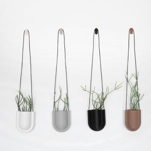 May include: Four hanging planters in white, grey, black, and brown, each with a black cord and a small plant. The planters are mounted on a white wall, creating a minimalist and modern aesthetic. The plants have thin, green stems.