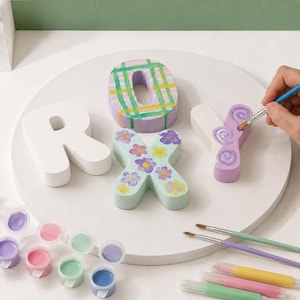 Peut inclure: Lettres en bois épelant "ROXY" peintes dans différentes couleurs et motifs. Le "O" est lavande avec un motif à carreaux vert et jaune. Le "X" est vert clair avec des fleurs violettes et jaunes. Des pots de peinture et des pinceaux sont également visibles.
