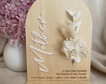 May include: A wooden arch with the word "Mother" in white lettering. A small bouquet of dried white flowers is attached to the arch. The text "A love that exceeds the beauty of any flower" is written below the flowers. The text "Love Alexis, Amari, Lalkin, Lotti, Blair & Bodhi" is written below the quote.
